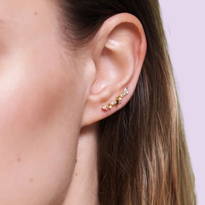 A close-up of a woman's ear wearing a small gold ear climber and crawler earrings adorned with tiny clear stones, set against a soft light purple background.
