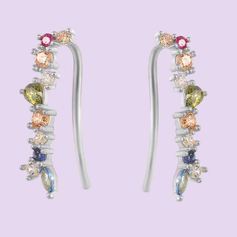 A pair of silver-toned ear crawlers and climber earrings with multiple colorful gemstones, including pink, yellow, blue, and peach-colored stones, set against a solid light purple background.