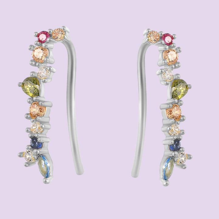 A pair of silver-toned ear crawlers and climber earrings with multiple colorful gemstones, including pink, yellow, blue, and peach-colored stones, set against a solid light purple background.