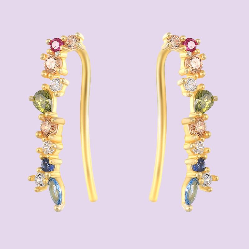A pair of gold ear crawler or climber earrings decorated with a vertical arrangement of colorful gemstones, including green, blue, pink, and clear crystals, set against a solid lavender background.