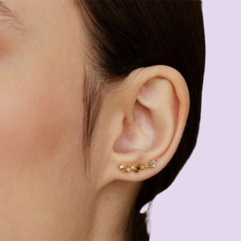 A close-up of a person's ear wearing a small cluster stud ear climber earrings or ear crawlers with gold and yellow-gold stones on a light lavender background.