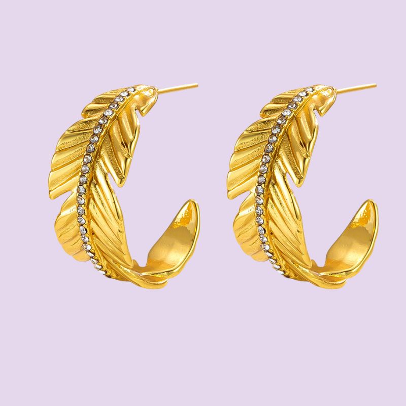 A pair of gold feather-shaped hoop earrings with a row of small clear rhinestones, displayed on a light lavender background.
