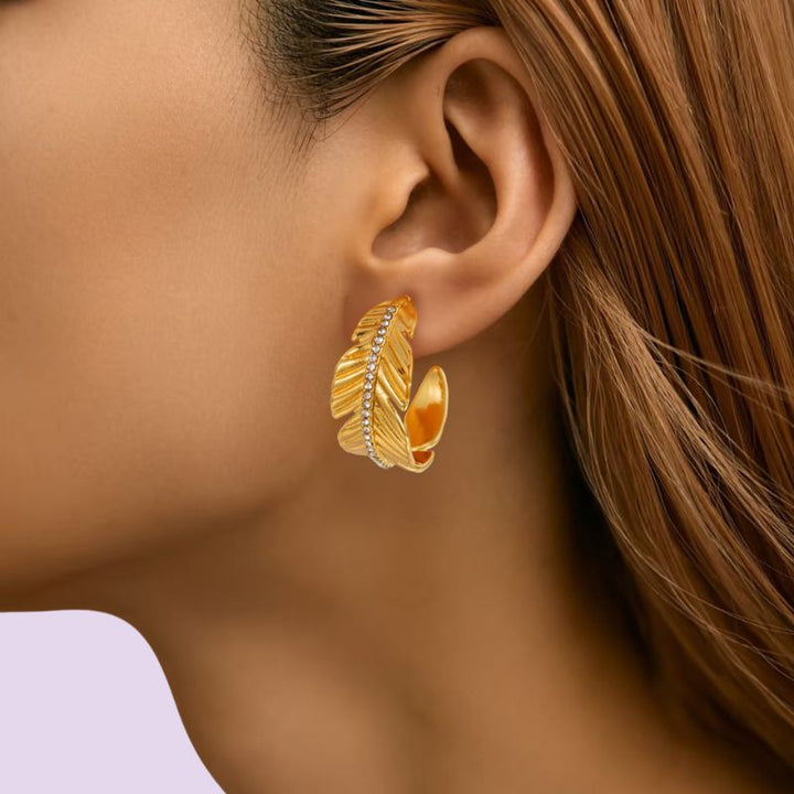 A close-up of a woman's ear wearing a gold huggie hoop earring with a textured feather design and a row of small clear stones.