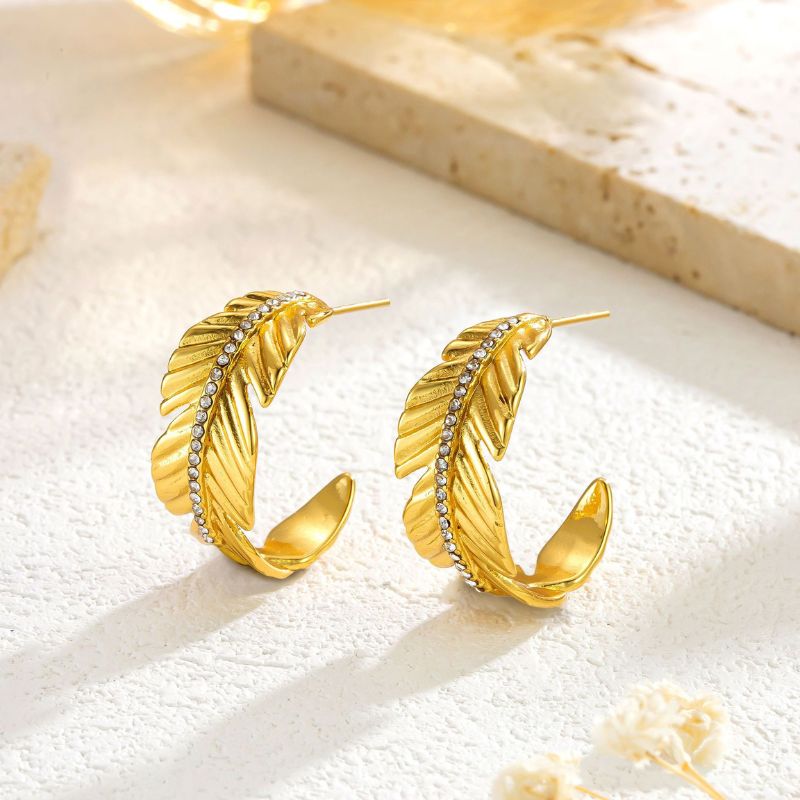 A pair of gold crescent-shaped hoop earrings designed as stylized feathers, accented with a row of small clear rhinestones, displayed on a textured light beige surface.
