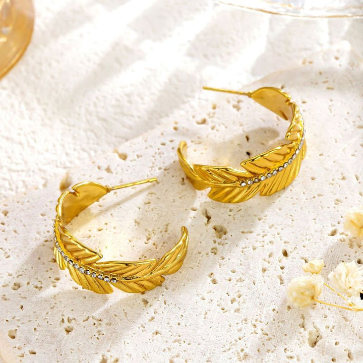 A pair of gold feather-shaped hoop earrings with small clear rhinestone accents, displayed on a textured white surface with dried flowers.