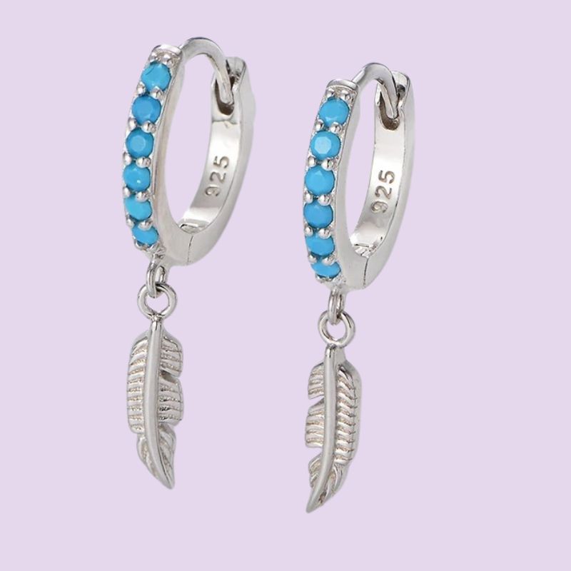 A pair of sterling silver huggie hoop earrings with a row of small round turquoise blue stones and a dangling textured feather charm, shown on a light lavender background.