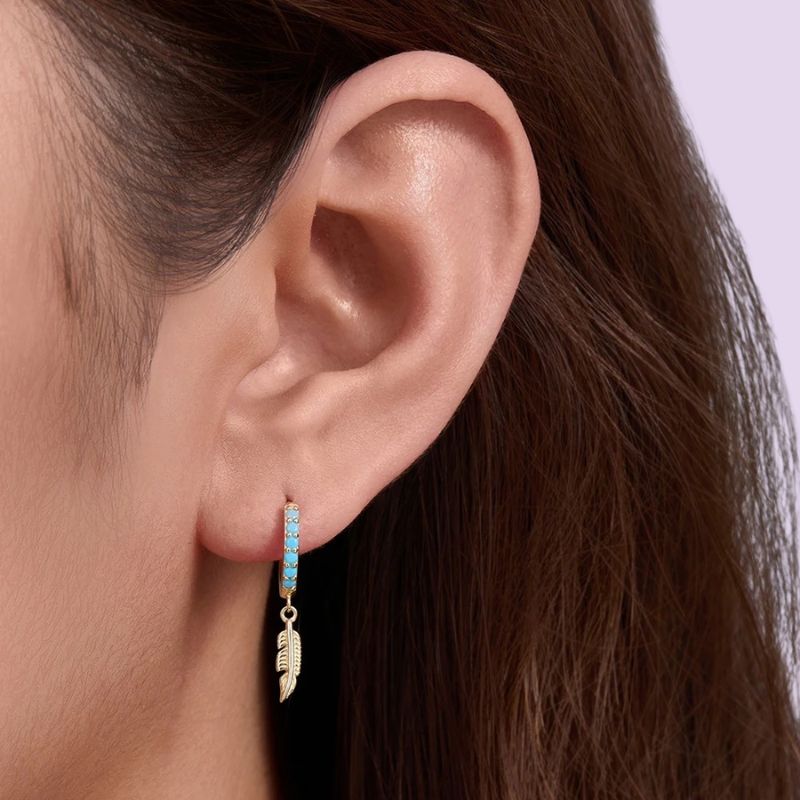 A close-up of a person's ear wearing a small gold hoop earring with a dangling feather charm and a row of light blue stones.