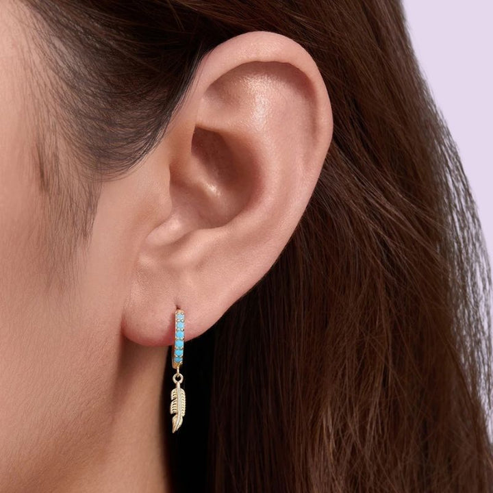 A close-up of a person's ear wearing a small gold hoop earring with a dangling feather charm and a row of light blue stones.