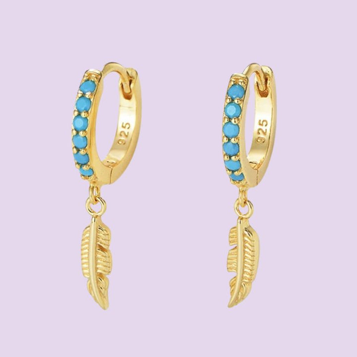 A pair of gold huggie hoop earrings with a row of turquoise blue stones and a dangling gold feather charm, shown on a light lavender background.