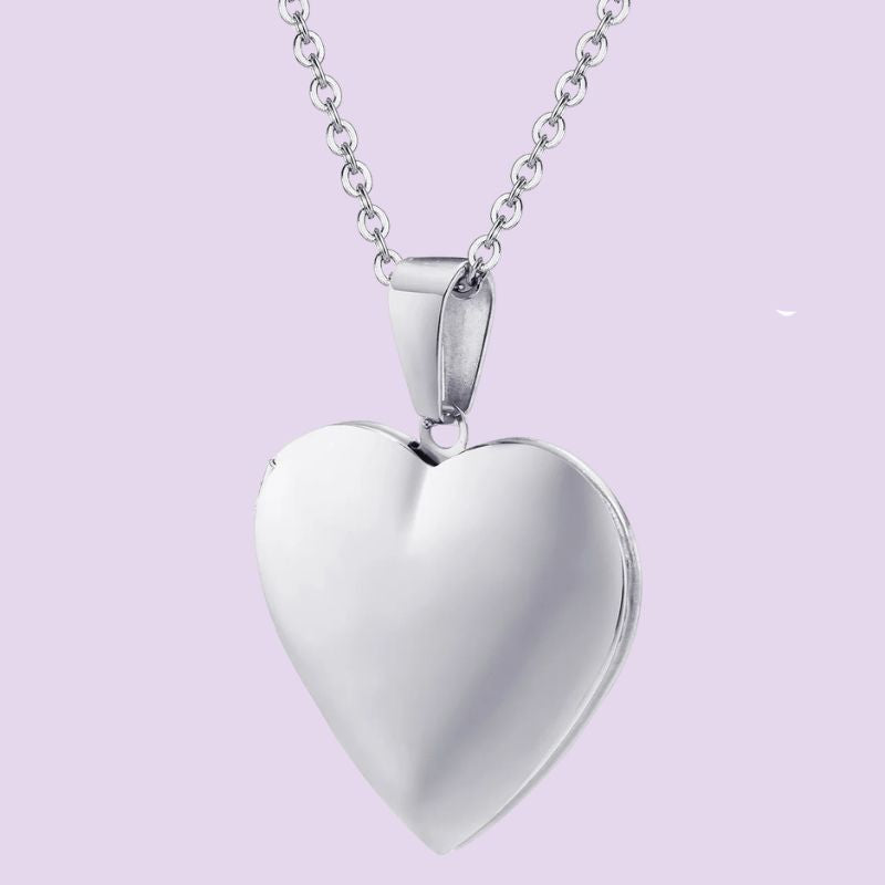 A polished silver heart-shaped locket pendant necklace on a matching silver chain, set against a solid light lavender background.