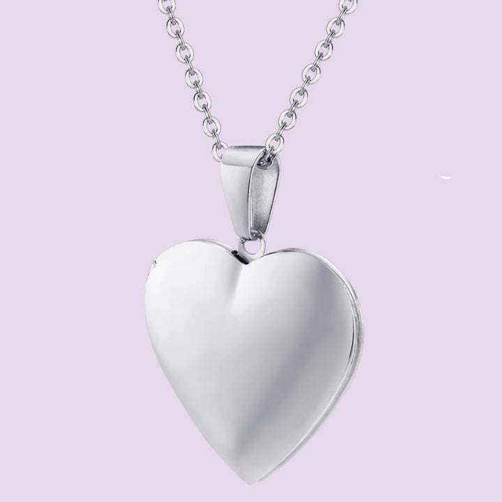 A polished silver heart-shaped locket pendant necklace on a matching silver chain, set against a solid light lavender background.
