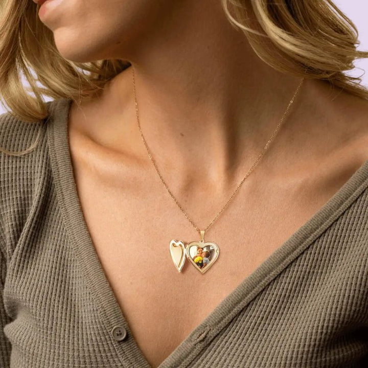 A close-up of a woman wearing a delicate gold locket necklace with two interlocking heart-shaped pendants, one of which contains a small printed photo, paired with a brown waffle-knit top.