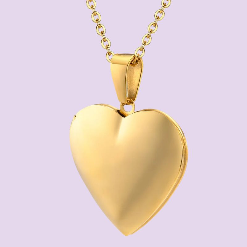 A polished yellow gold heart-shaped locket pendant necklace on a matching chain, set against a solid light lavender background.