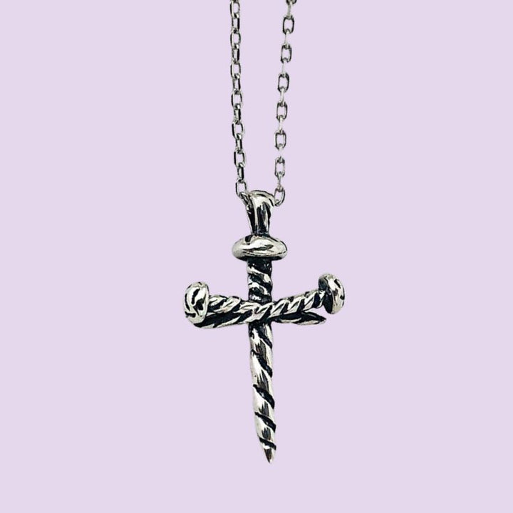 A silver-toned cross nail pendant necklace with a twisted rope design , hanging from a matching chain against a solid light purple background.