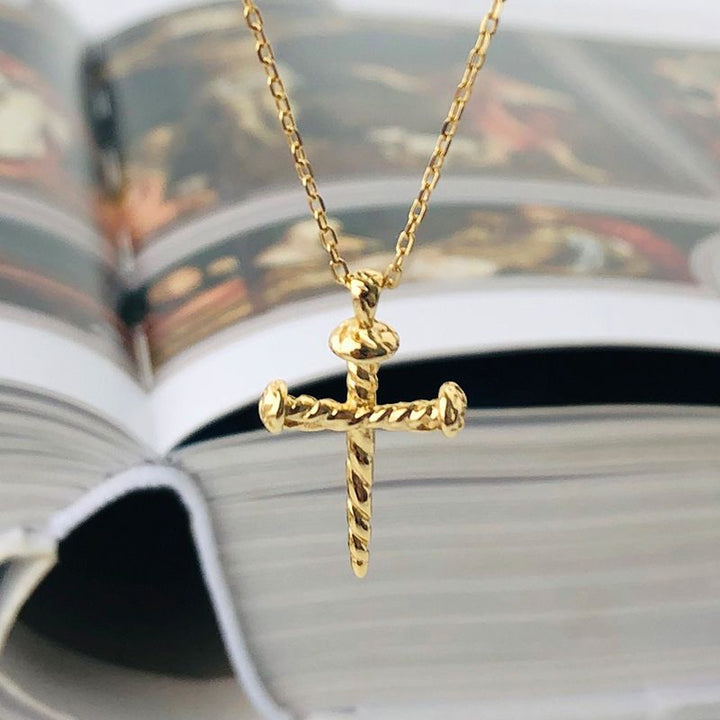 A gold cross nail pendant necklace with a twisted rope design hangs over an open book. 