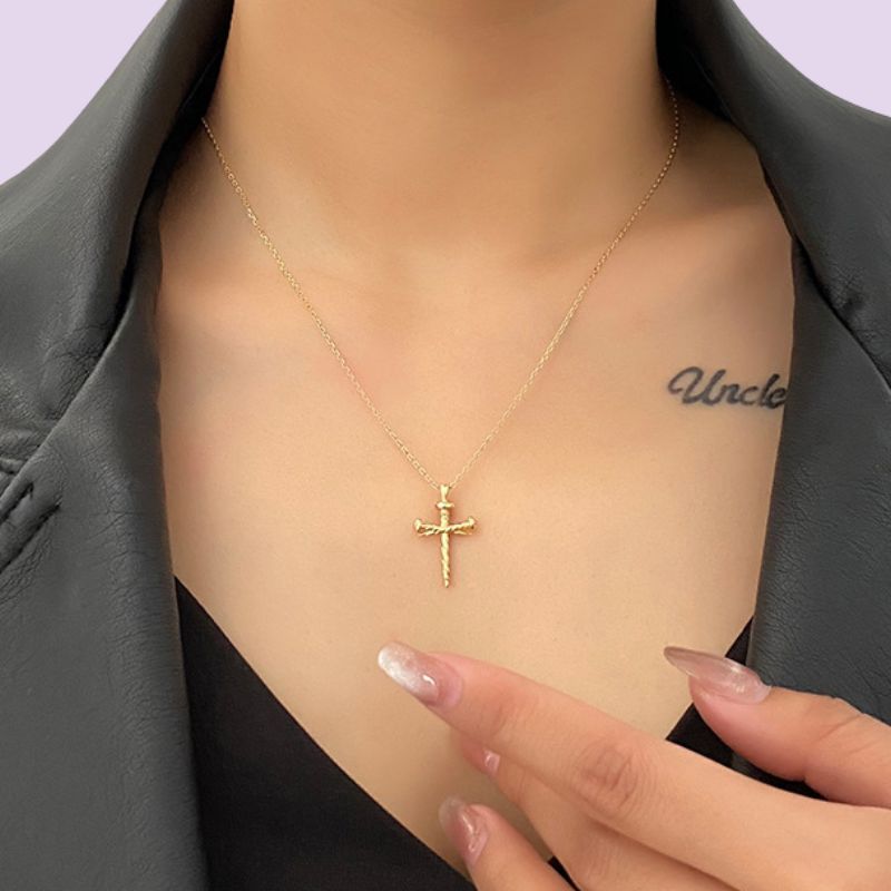A close-up of a model wearing a delicate gold cross nail pendant necklace, styled with a black top and dark gray blazer. The model's hand with manicured nails rests near the necklace.
