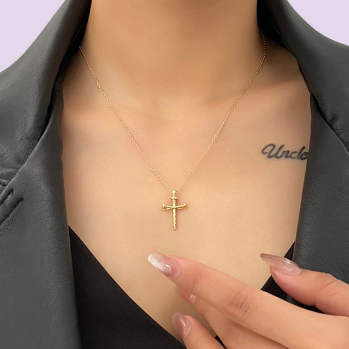 A close-up of a model wearing a delicate gold cross nail pendant necklace, styled with a black top and dark gray blazer. The model's hand with manicured nails rests near the necklace.