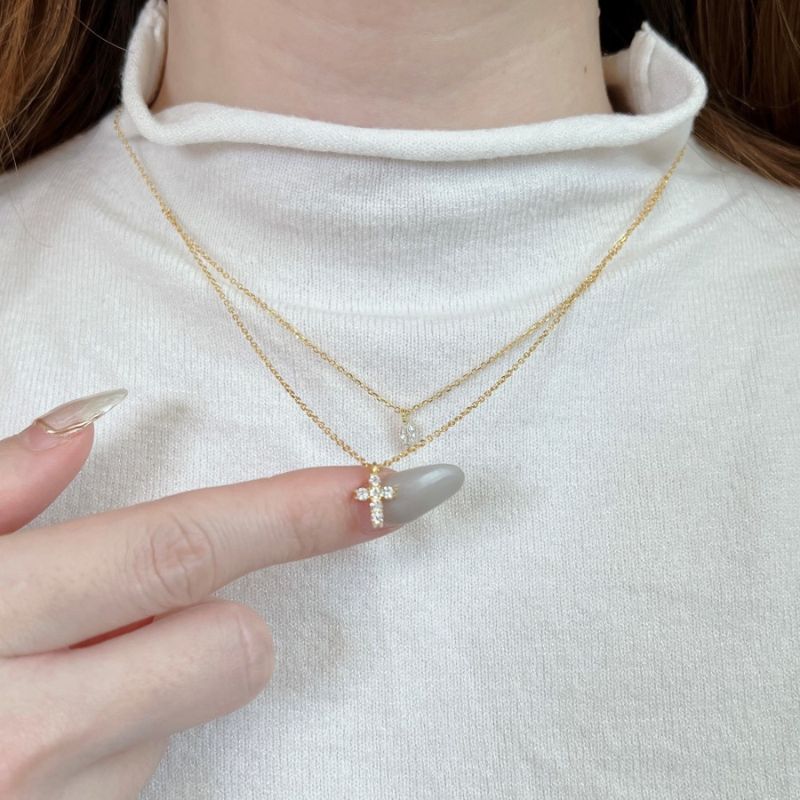A close-up of a person wearing a delicate double-layered gold necklace with a small cross pendant adorned with clear crystals, styled with a white ribbed turtleneck sweater.