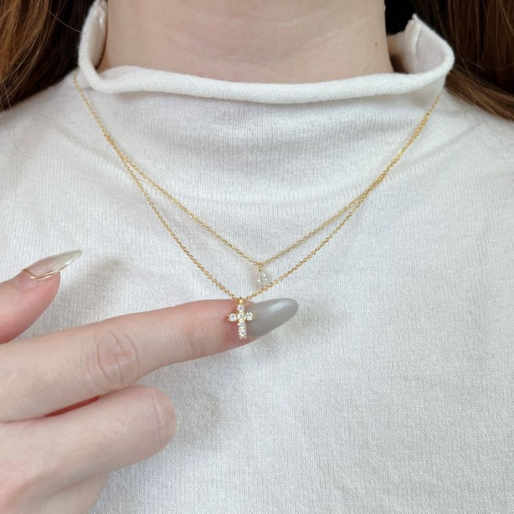 A close-up of a person wearing a delicate double-layered gold necklace with a small cross pendant adorned with clear crystals, styled with a white ribbed turtleneck sweater.