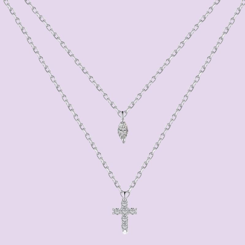 A silver double-layer necklace featuring a delicate chain with a small teardrop-shaped crystal pendant on the top layer and a thicker chain with a cross pendant encrusted with clear crystals on the bottom layer, set against a solid light purple background.