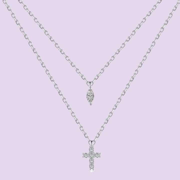 A silver double-layer necklace featuring a delicate chain with a small teardrop-shaped crystal pendant on the top layer and a thicker chain with a cross pendant encrusted with clear crystals on the bottom layer, set against a solid light purple background.