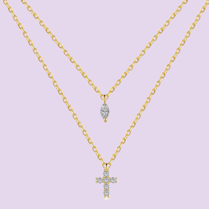A double-layered gold necklace set on a light lavender background. The top layer features a small teardrop-shaped pendant with clear stones, and the bottom layer features a larger cross pendant with a halo of clear stones.