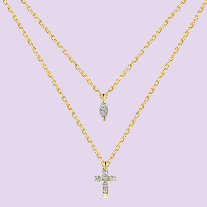 A double-layered gold necklace set on a light lavender background. The top layer features a small teardrop-shaped pendant with clear stones, and the bottom layer features a larger cross pendant with a halo of clear stones.
