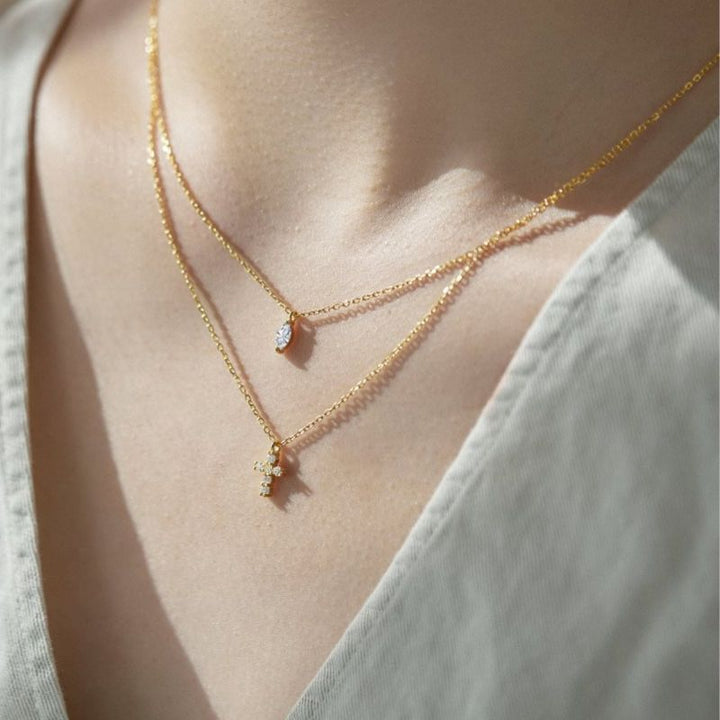 A close-up of a person's neck and chest wearing a delicate layered gold necklace with a small oval and a small cross charm, styled with a light gray V-neck top.