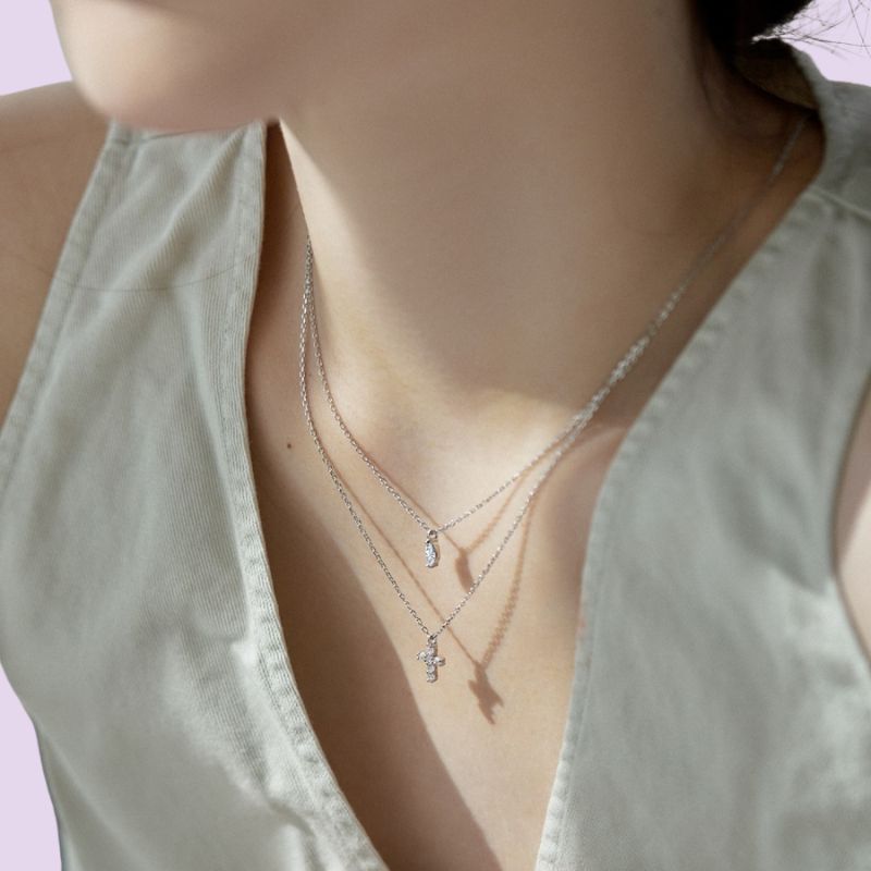 A close-up of a model wearing a layered silver necklace set with a small crescent moon and cross pendants, styled with a light sage green sleeveless top.